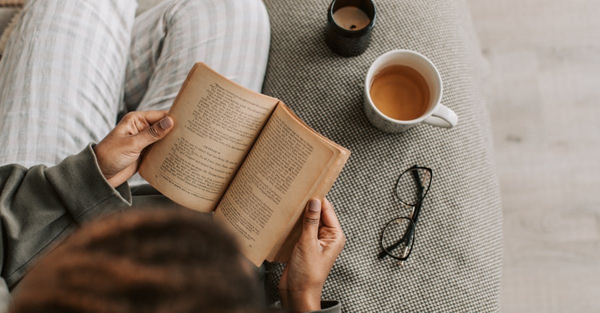 A woman is sitting on a couch reading a book and drinking coffee.