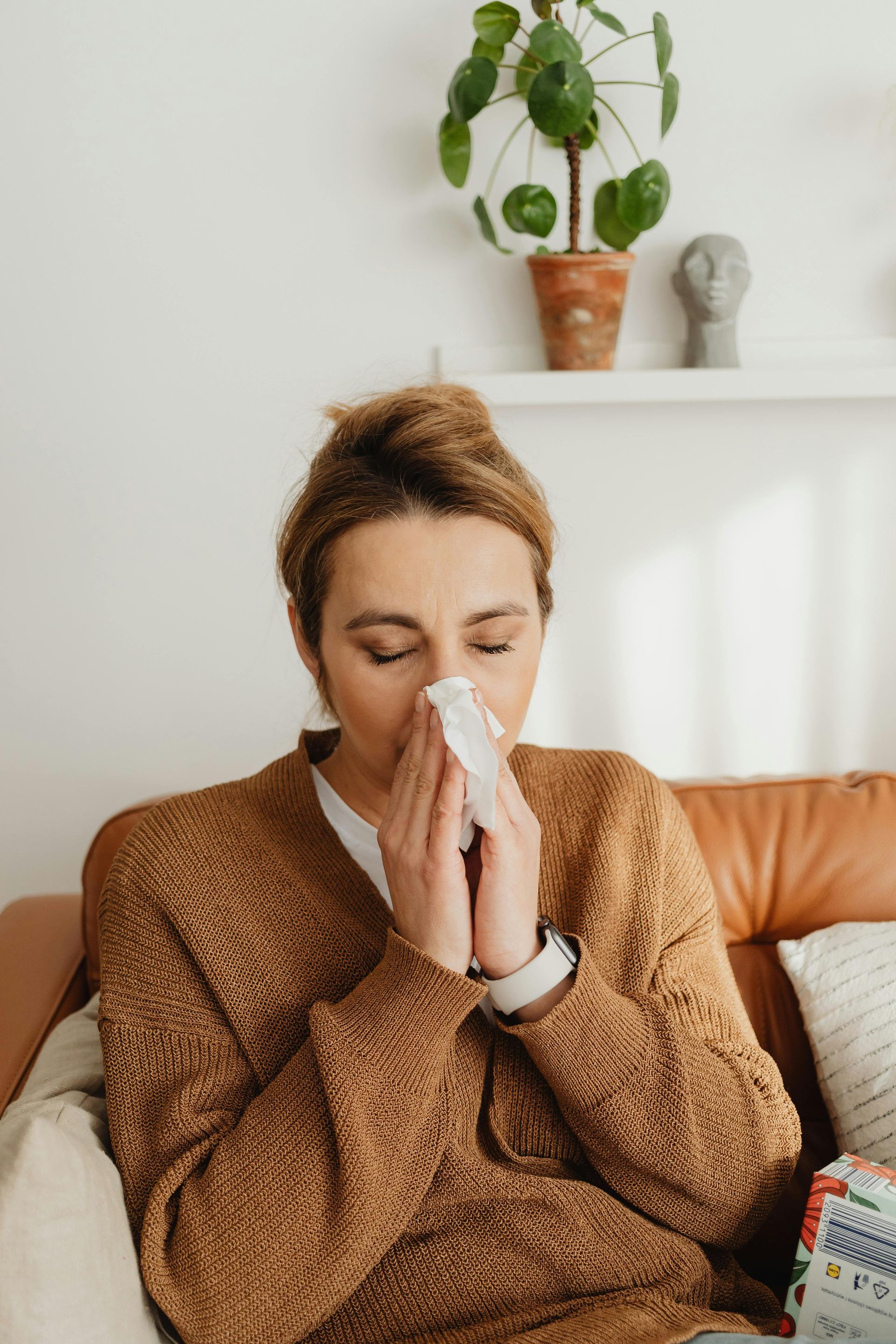 Woman seated on a couch, blowing her nose with a tissue; brown sweater, home setting.