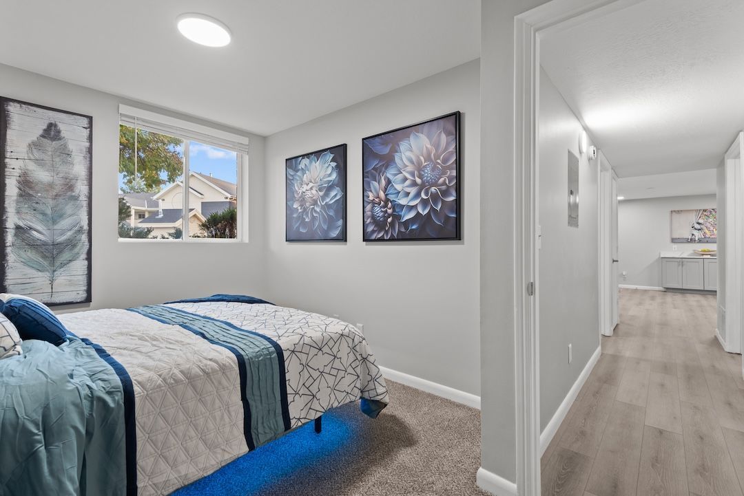 Bright bedroom with blue bedding, framed wall art, and a hallway leading to a kitchen area.