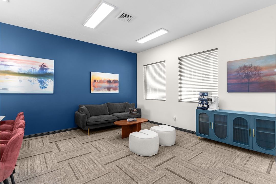 Modern lounge with blue accent wall, black sofa, white stools, and framed art in a bright office space