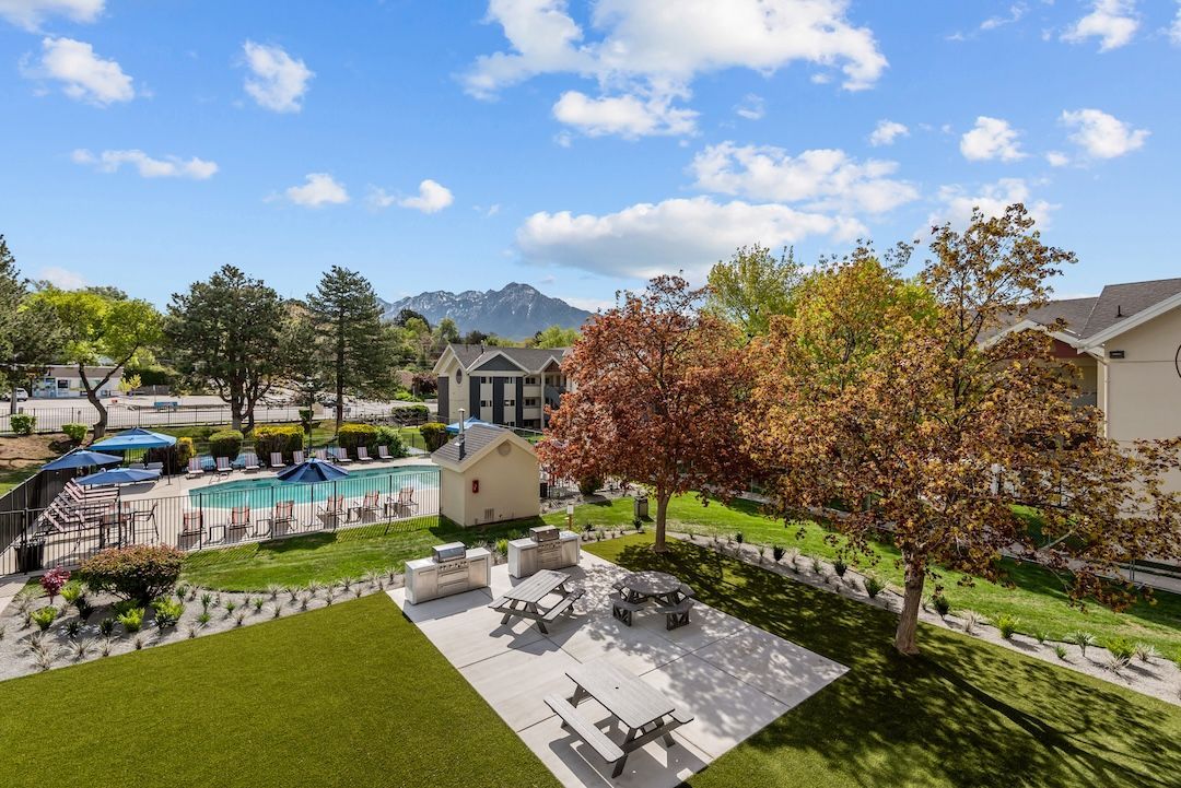 Apartment courtyard with pool, patio seating, green lawn, and autumn trees under a blue sky