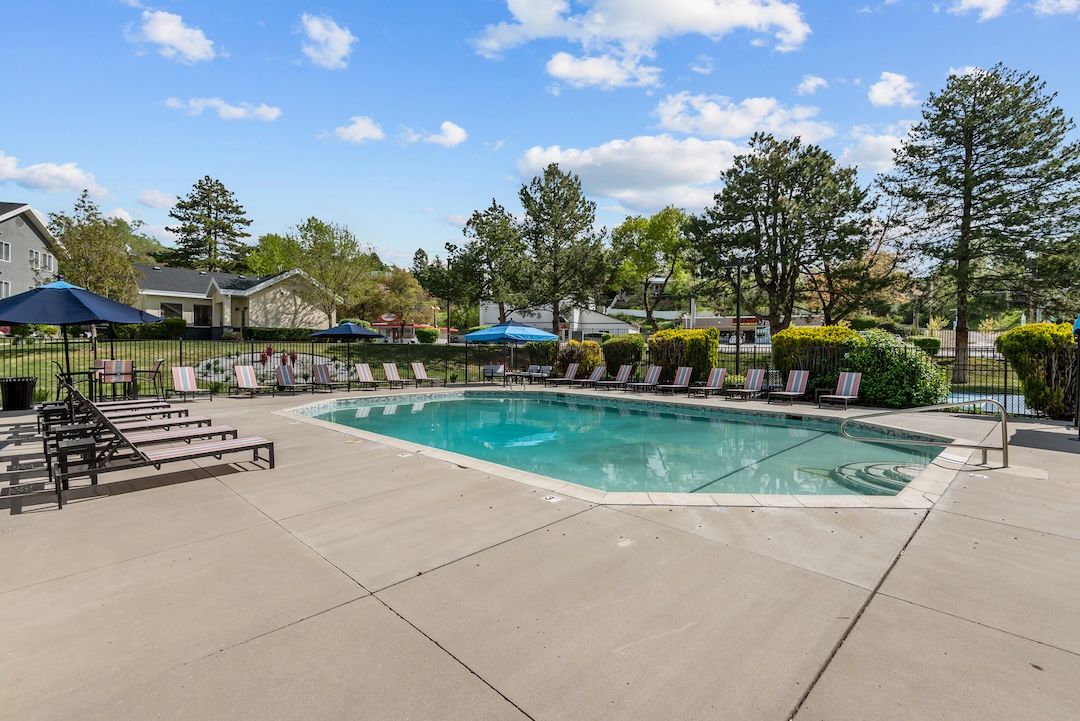Outdoor swimming pool with lounge chairs, patio umbrellas, and trees under a blue sky.