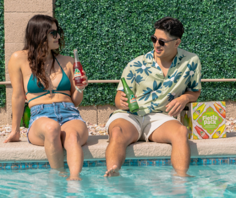 A man and a woman are sitting on the edge of a swimming pool holding drinks.