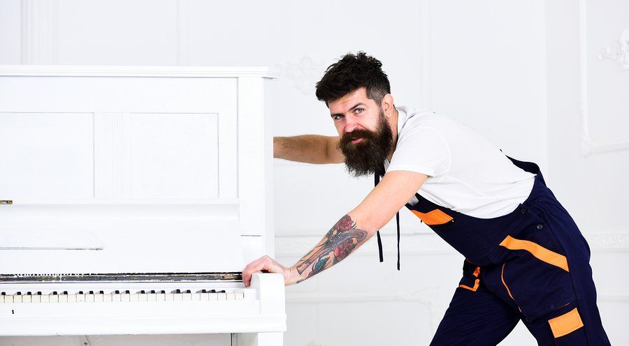 Man pushing a white piano; wearing navy and orange overalls, tattoo on arm. White room.