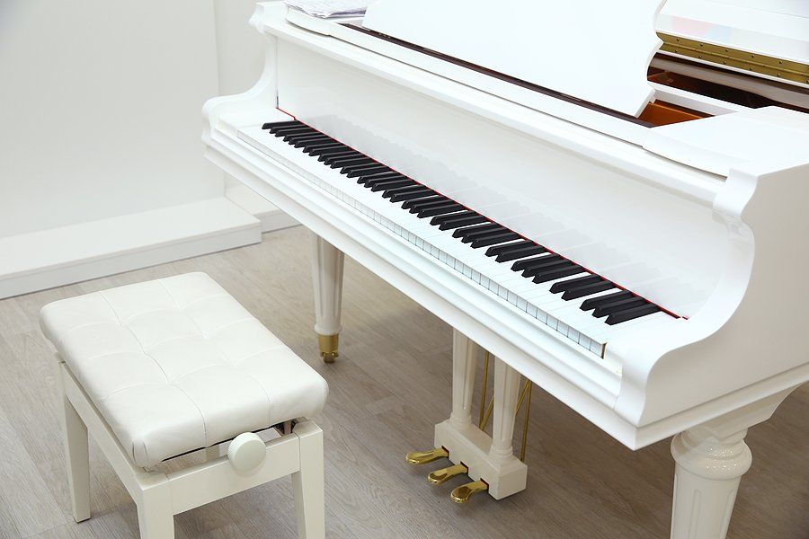 White grand piano with matching bench. Three brass pedals, black and white keys, in a white room.