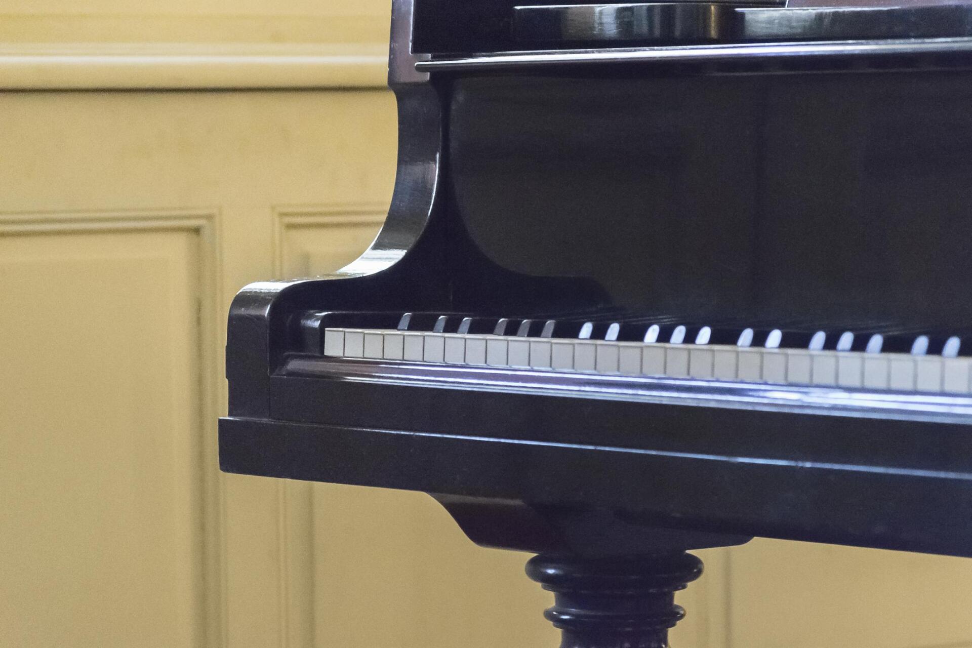 Black grand piano with white and black keys, against a light yellow paneled wall.
