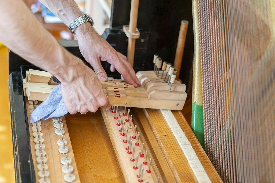 Person cleans a piano mechanism with a cloth. Wooden parts, metal pins, and strings are visible.