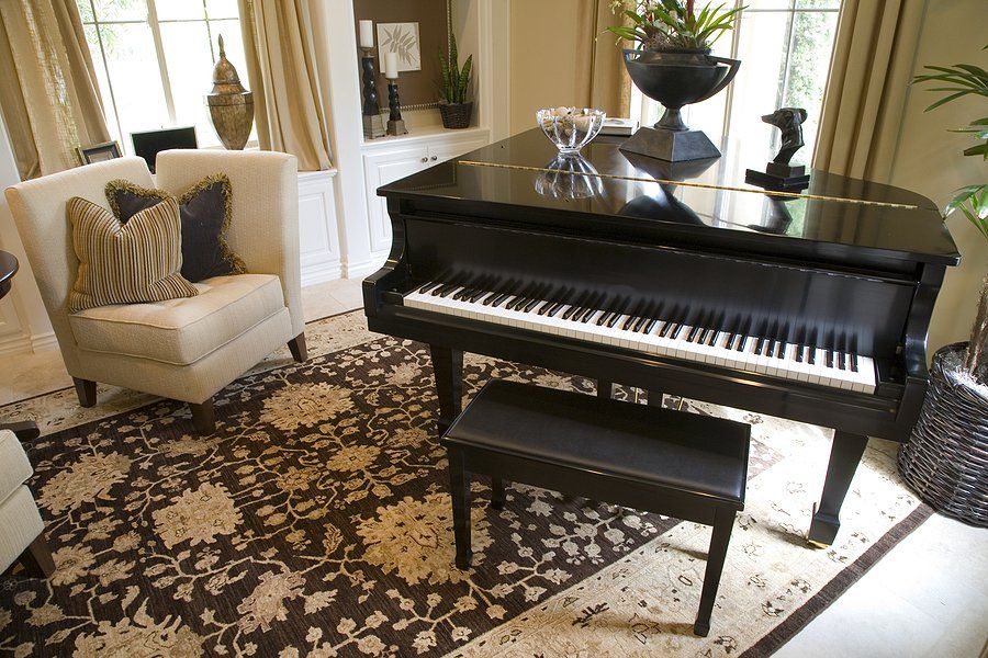 Black grand piano with open lid, set in a living room with a patterned rug and armchairs.