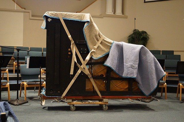Upright piano on a dolly, covered with moving blankets and secured with straps, in an auditorium.