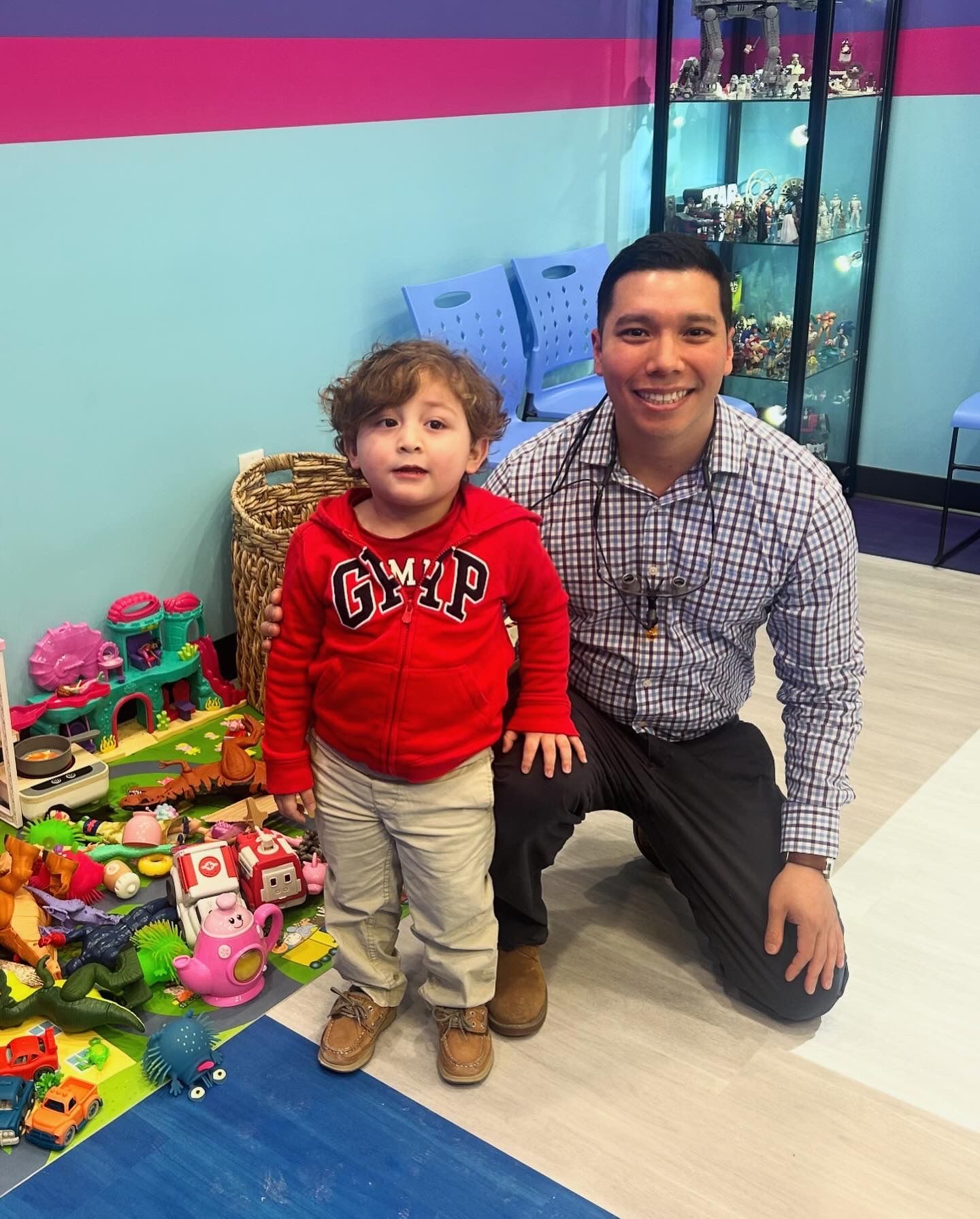 A smiling man kneels beside a child in a colorful playroom filled with toys.