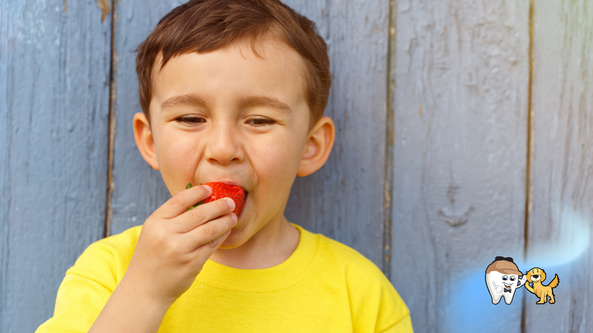 Boy in yellow shirt eating a red strawberry in front of blue wooden planks.