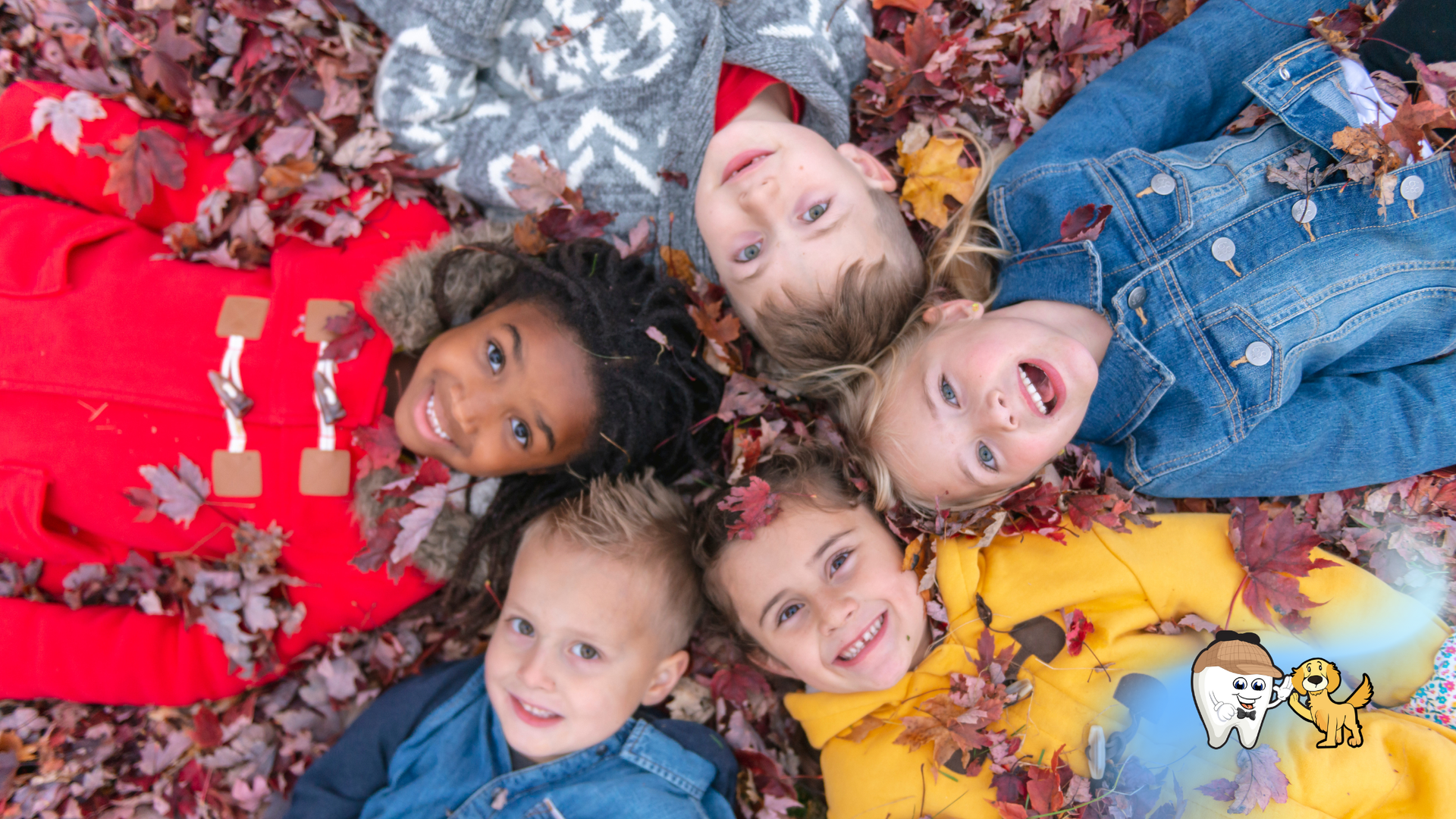 Children lying in a circle on fall leaves, looking up and smiling.
