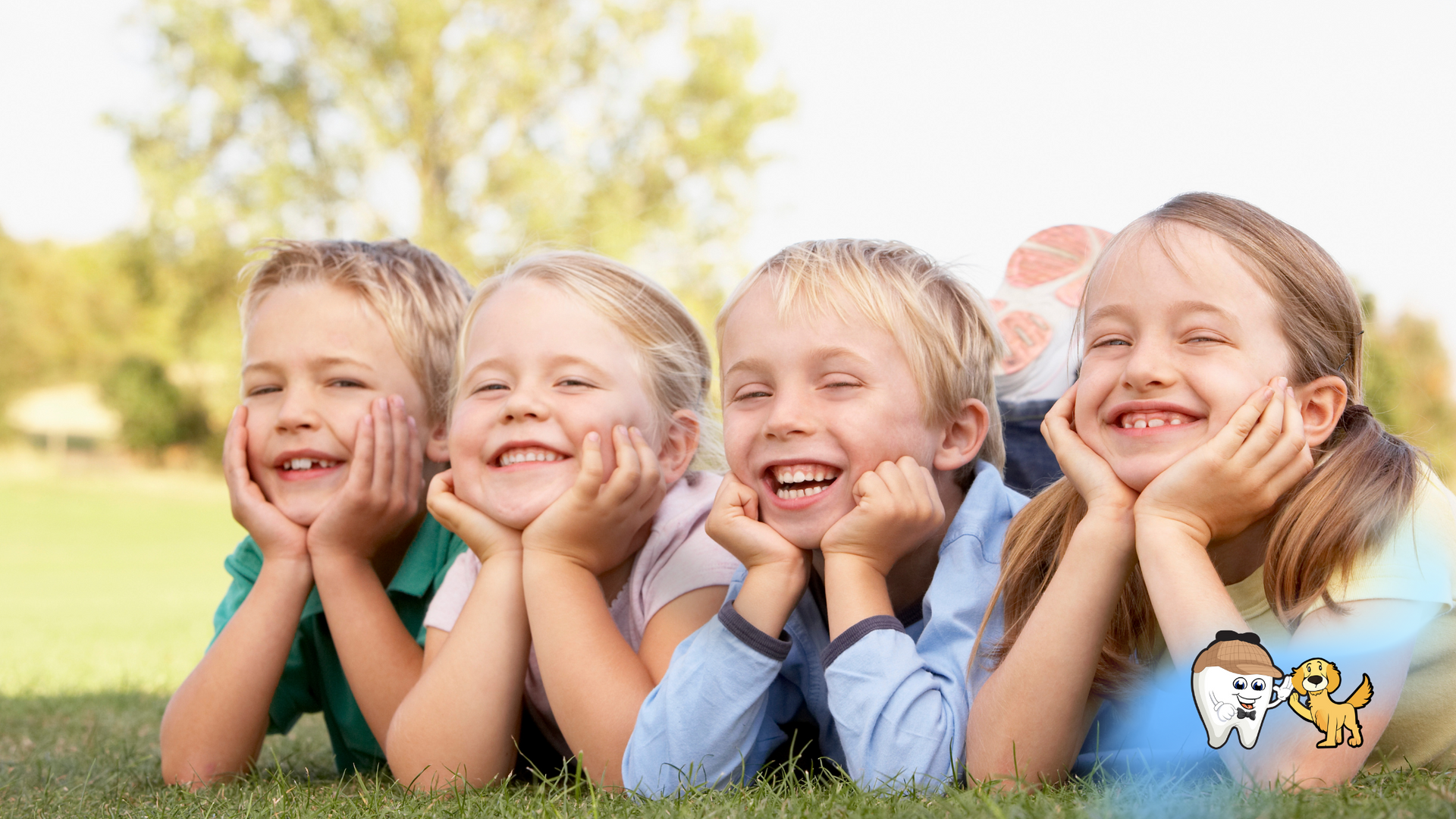 Four smiling children lying on grass, hands on chins, in a sunny outdoor setting.
