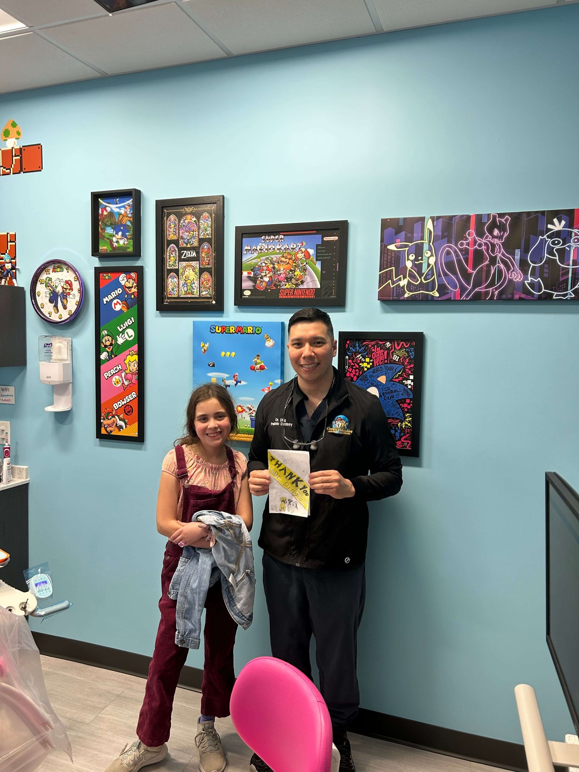 Dentist and young patient pose in a dental office, holding a paper; blue wall with art.