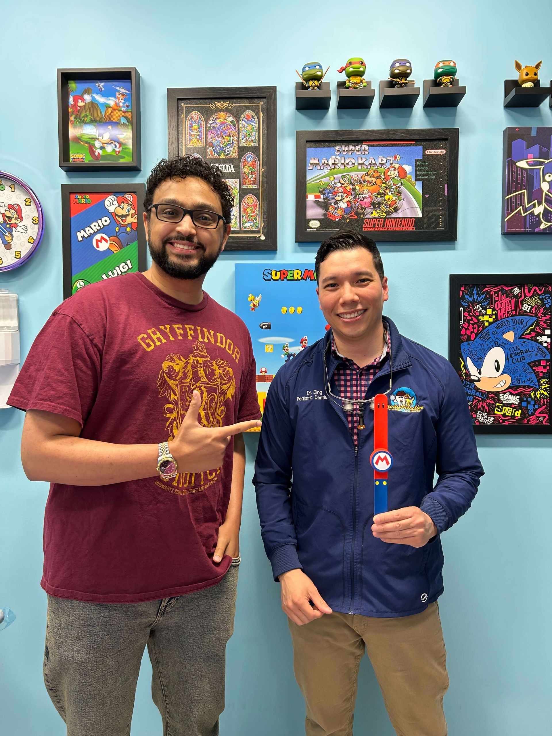 Two men pose with a Mario-themed item. They stand in front of a wall decorated with pixel art.