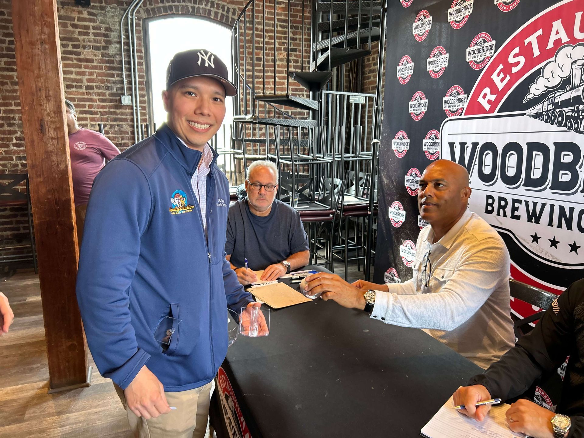 Man in a Yankees hat smiles, standing by a table where two men sign autographs at Woodbury Brewing.