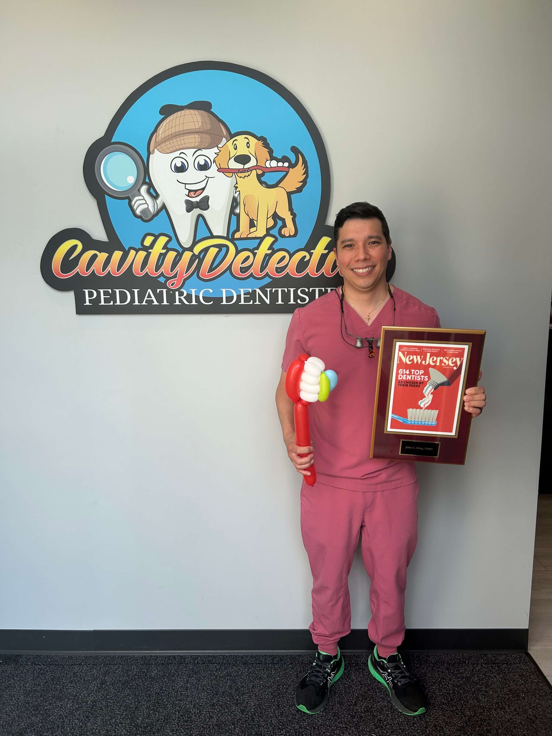 Man in pink scrubs holding a balloon and award plaque in front of a pediatric dentist office logo.