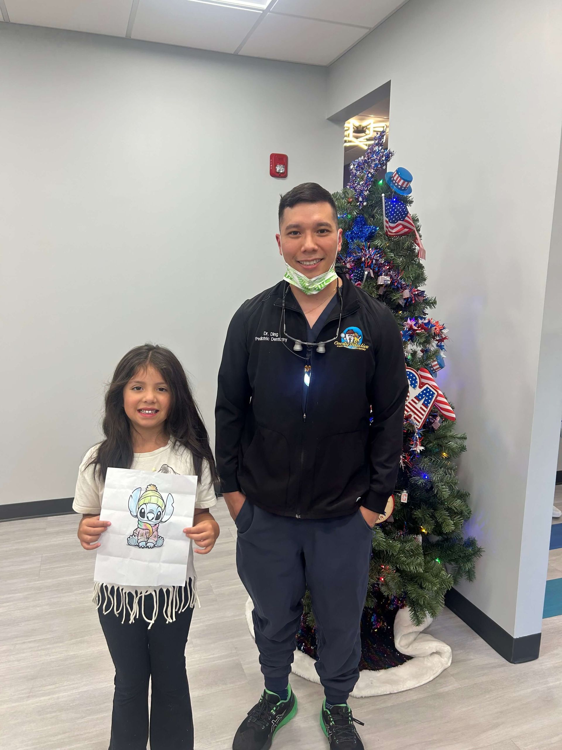 A child and a person in scrubs stand near a Christmas tree. The child holds a drawing.