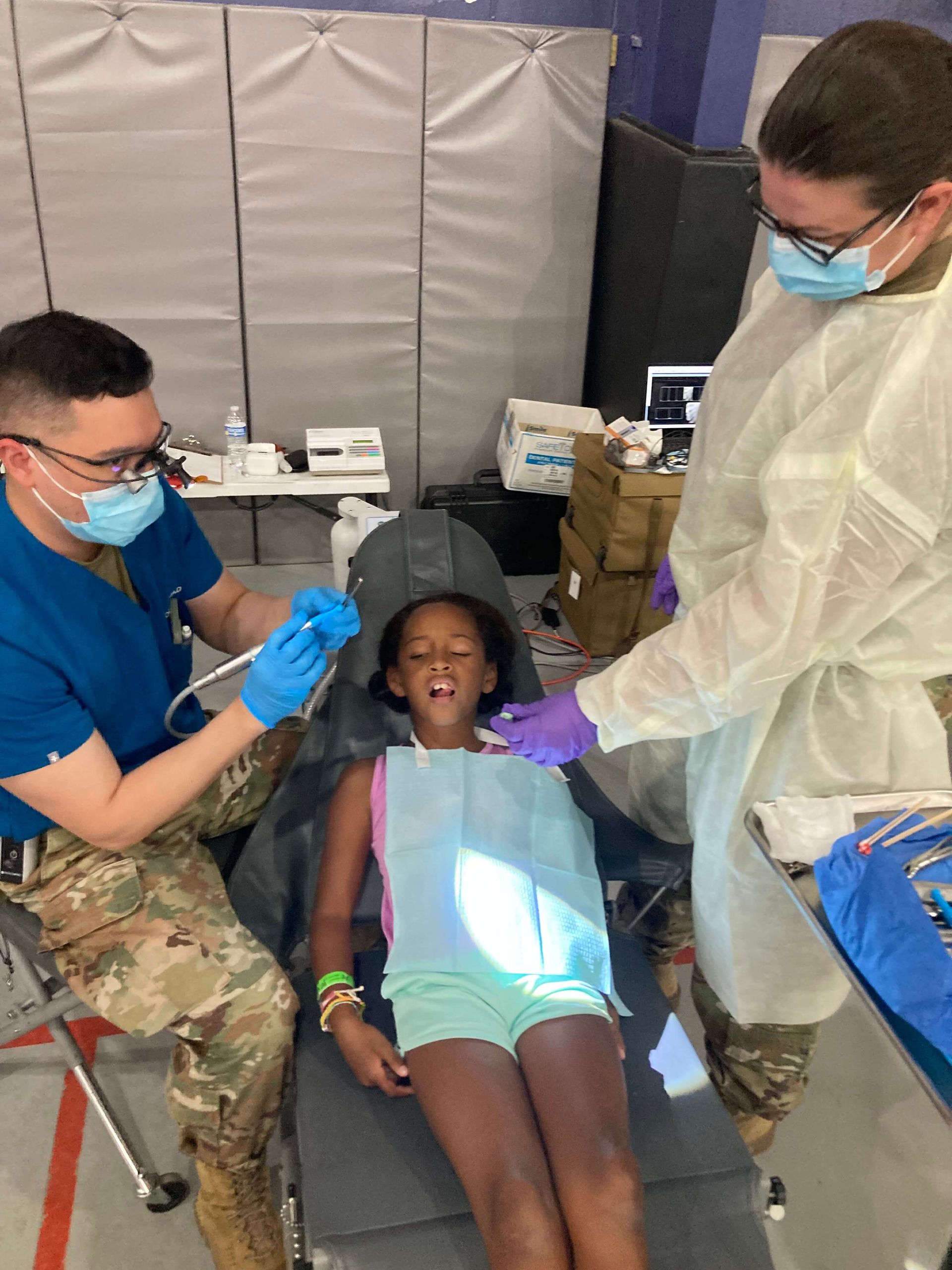 Child receiving dental care; two dental professionals in masks, one operating dental tools, clinic setting.
