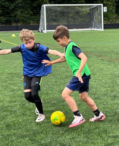 Two boys playing soccer on a green field; one in blue, one in green, kicking the yellow ball.