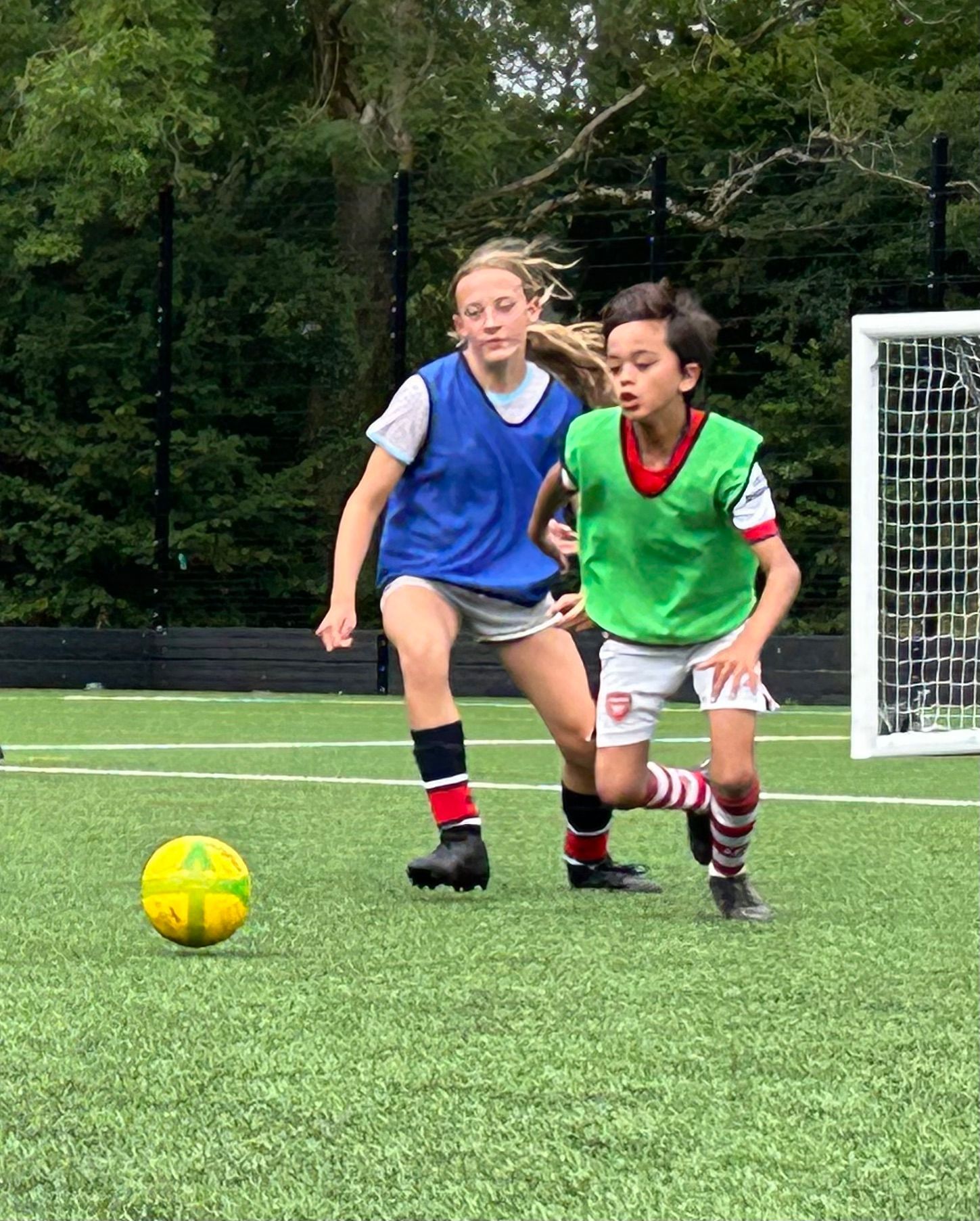 Two children playing soccer on green field. One wears blue vest, the other green. A yellow ball is on the grass.