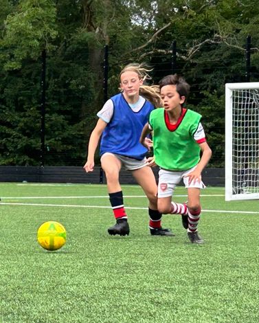 Two children playing soccer on green field. One wears blue vest, the other green. A yellow ball is on the grass.