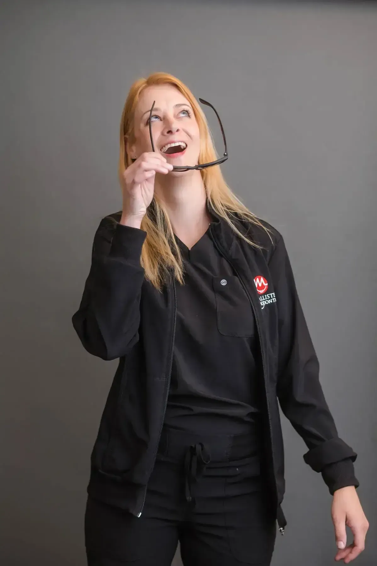 A woman in a black jacket is holding a pair of glasses over her mouth while laughing.