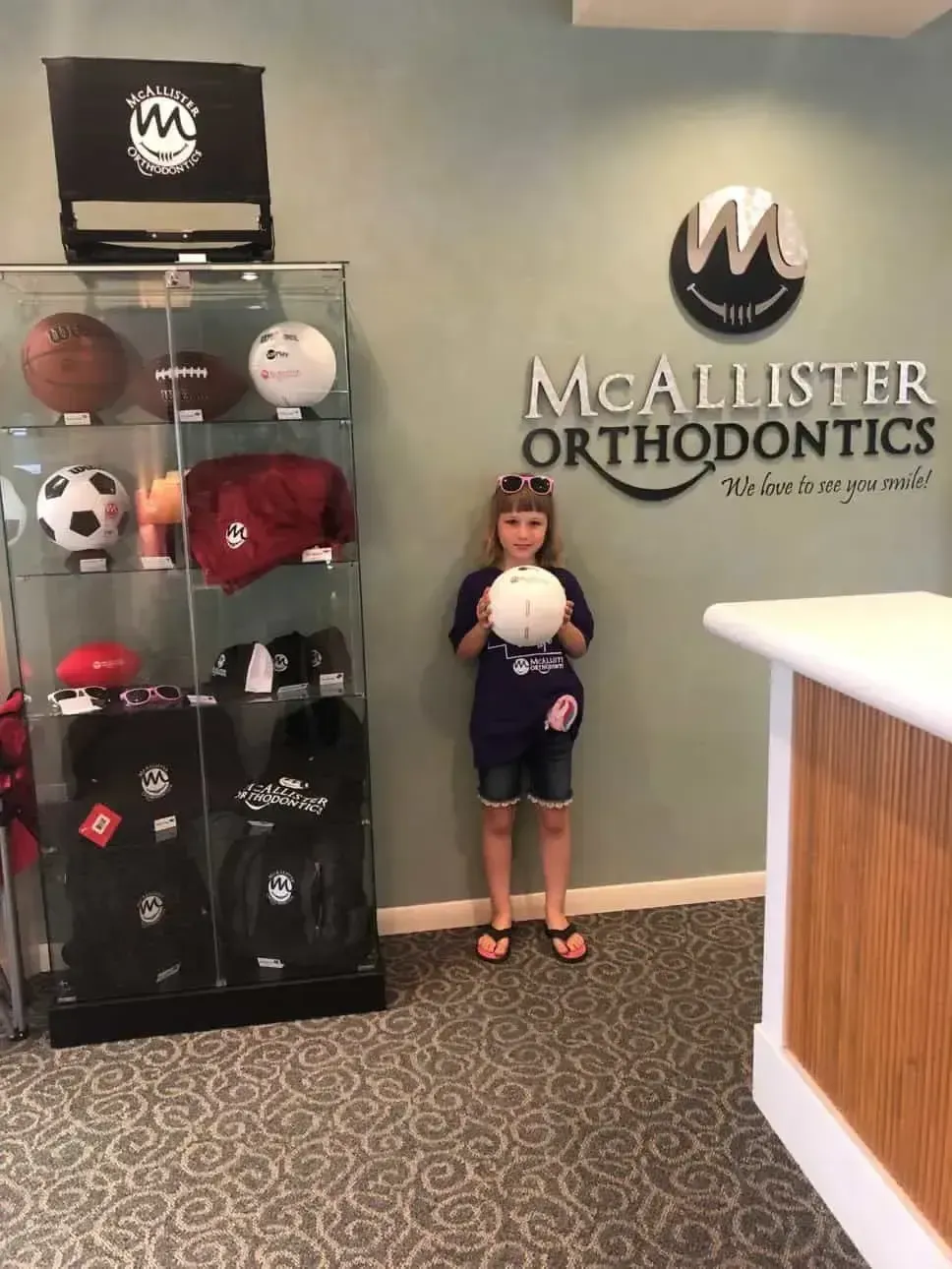 A little girl is holding a volleyball in front of a McAllister Orthodontics sign.