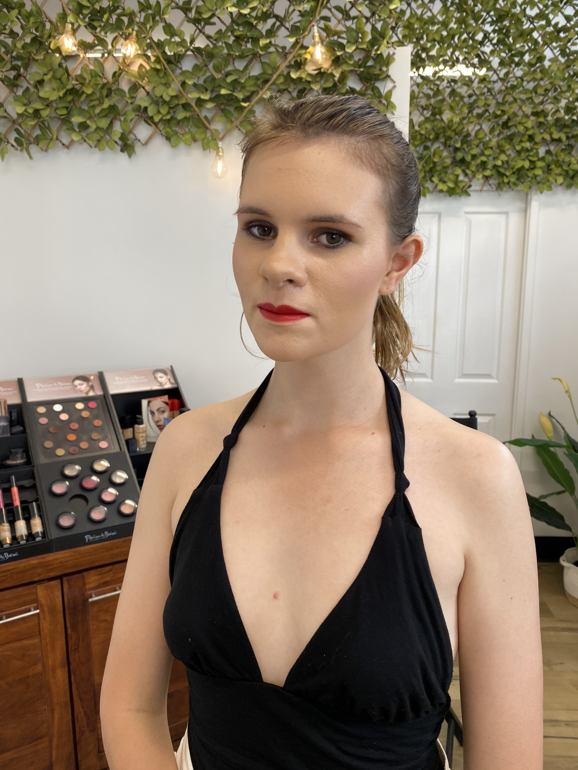 Person in a black halter dress standing indoors, with makeup display and greenery behind them — Penelope's Beauty in Raymond Terrace, NSW