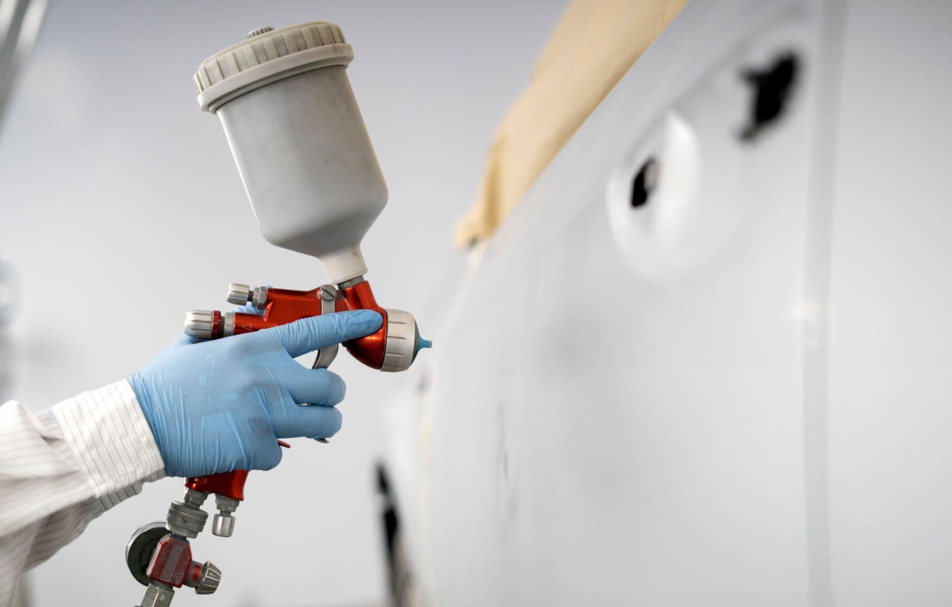 Person wearing a blue glove holding a red and gray paint sprayer, spraying a white car door in a garage.