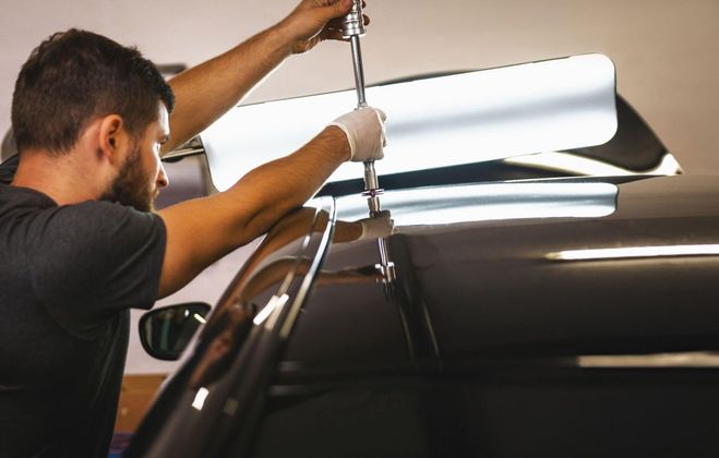 A man using a dent removal tool on a black car roof. He wears gloves and works under a bright light.
