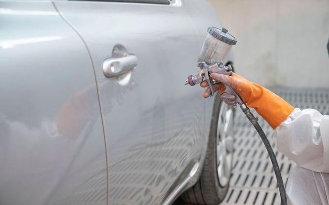 Person in protective gear spray-painting a silver car door with a paint sprayer in a workshop.