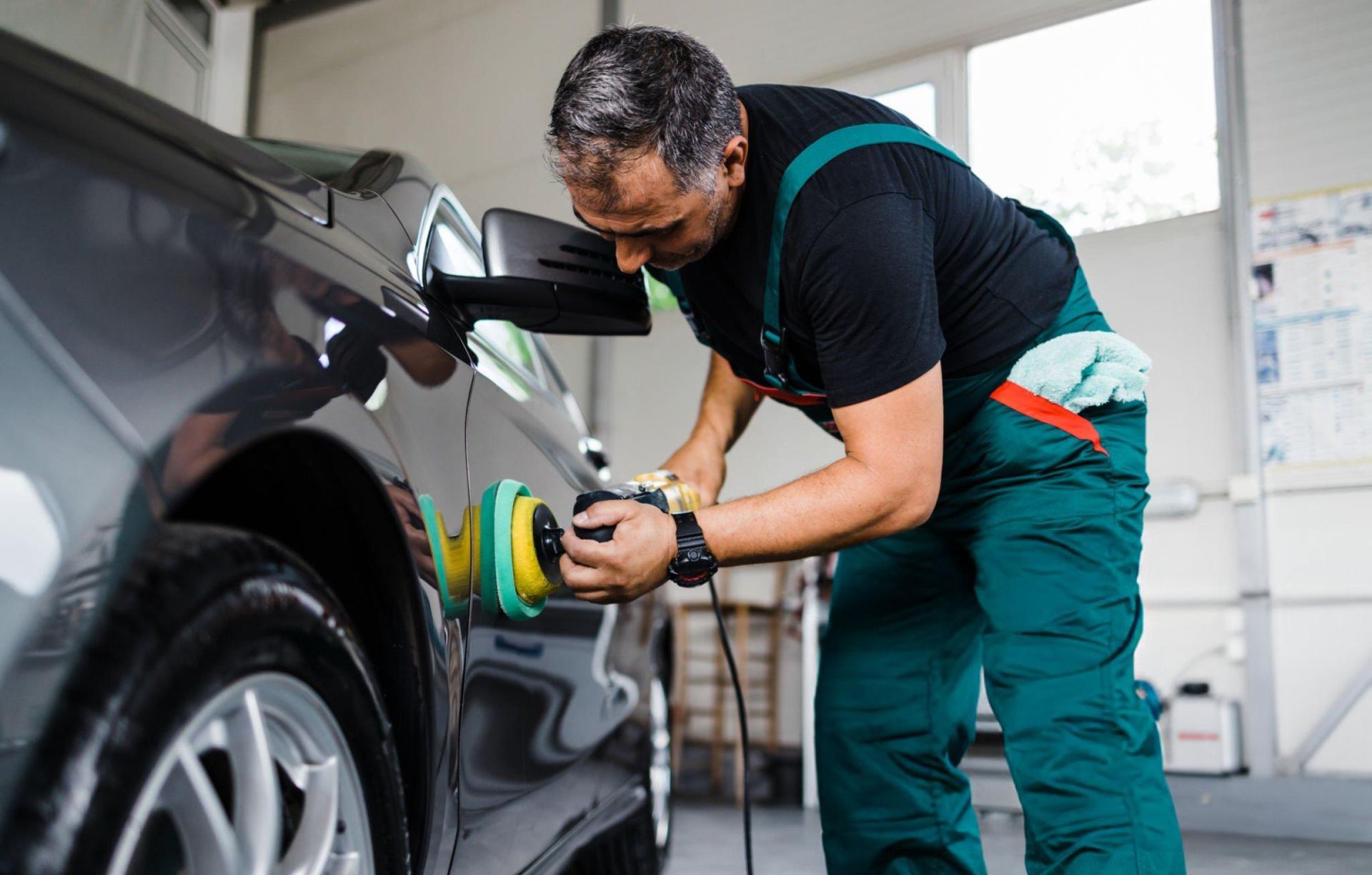 Man polishing a gray car with a machine polisher in a well-lit garage setting. He wears green overalls and a black shirt.