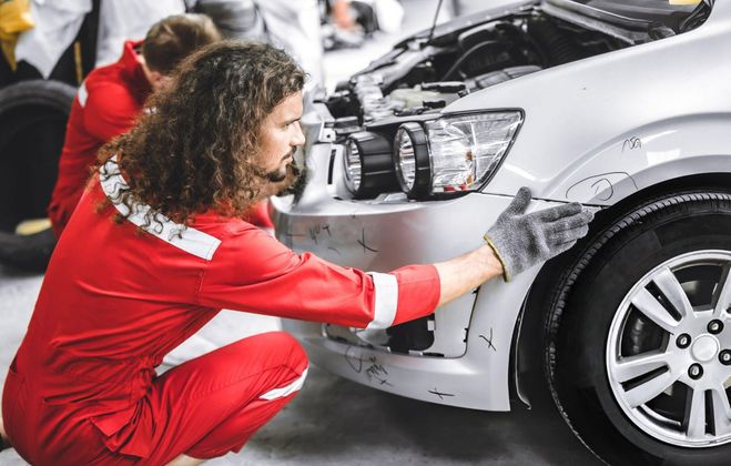 Mechanic in red overalls inspects a damaged silver car bumper, assessing scratches and dents.