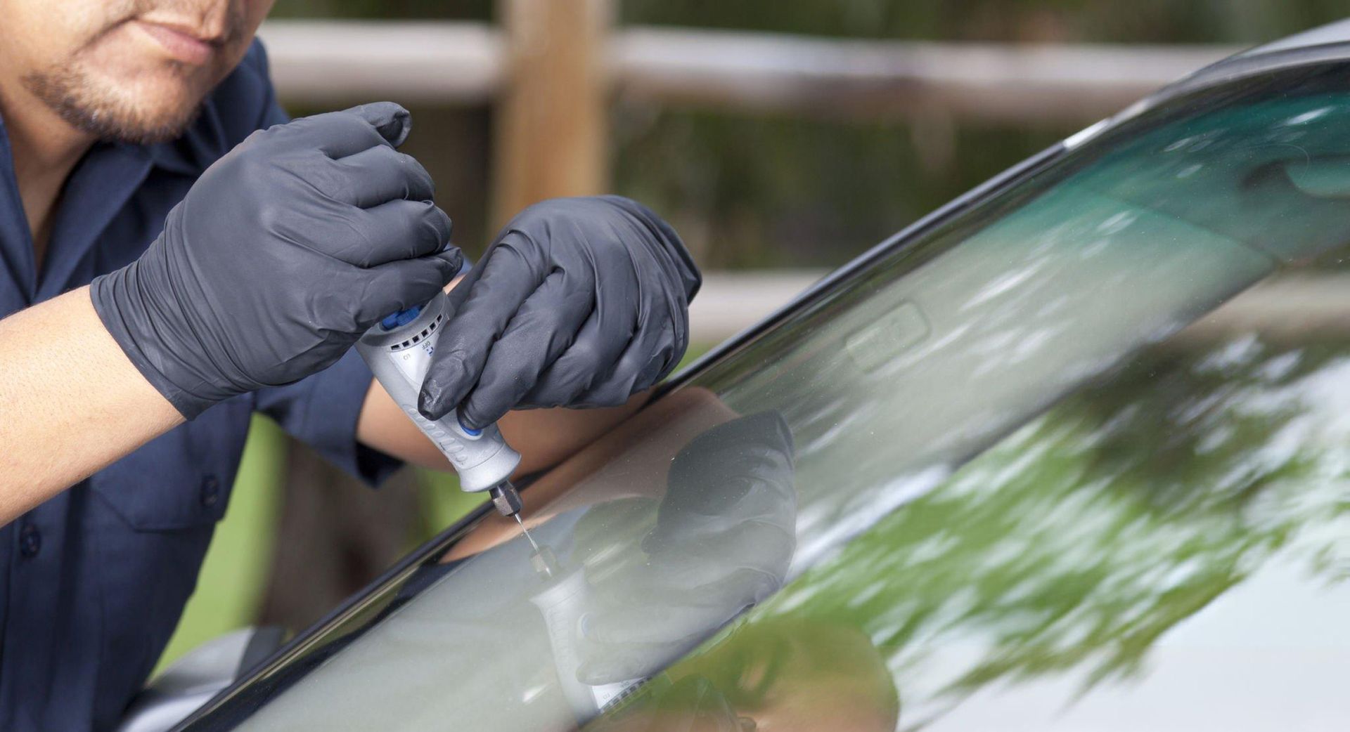 Person wearing black gloves repairing a windshield crack with a syringe, outdoors.