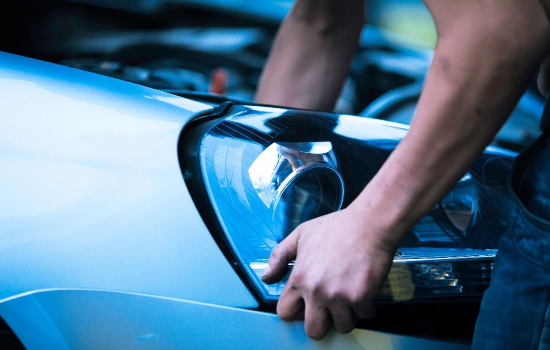 Person working on a car, leaning over and adjusting a headlight; blue tones.