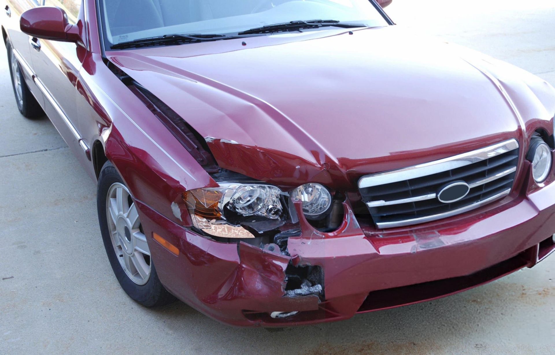 Damaged maroon car with crumpled hood and missing headlight, parked on a concrete surface.