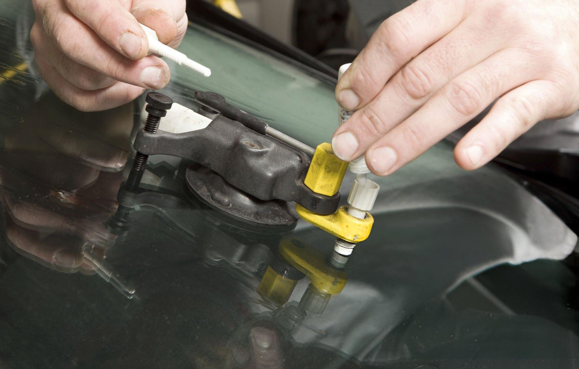 Hands using a repair kit to fix a crack in a car windshield. The kit is black and yellow, and the windshield is green.