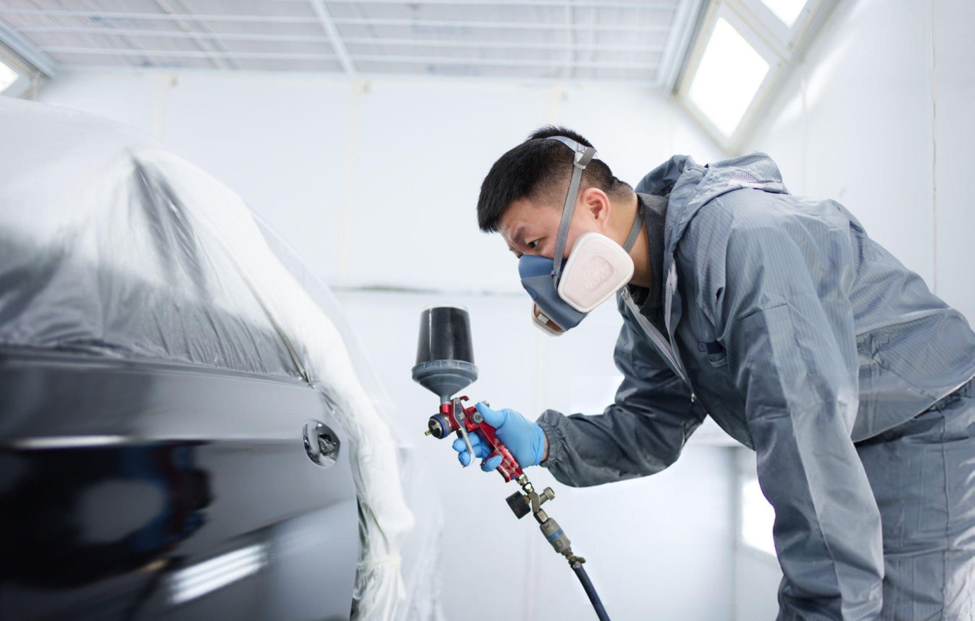 Man in respirator paints car in a spray booth. He wears a gray coverall and holds a spray gun.
