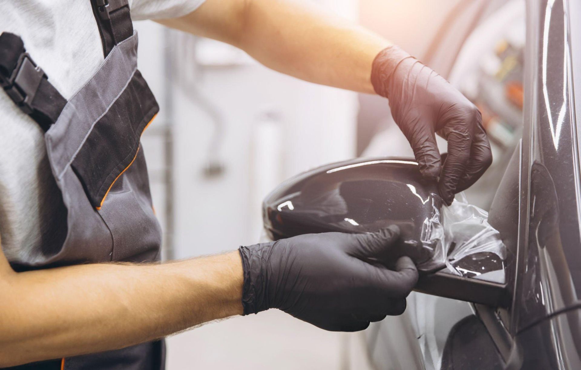 Mechanic in black gloves installing protective film on a car's side mirror.