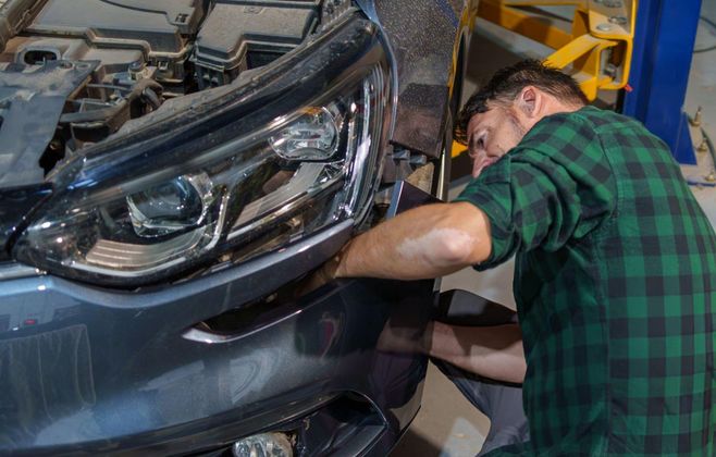 Mechanic in a green plaid shirt working on a car's headlight in a repair shop.