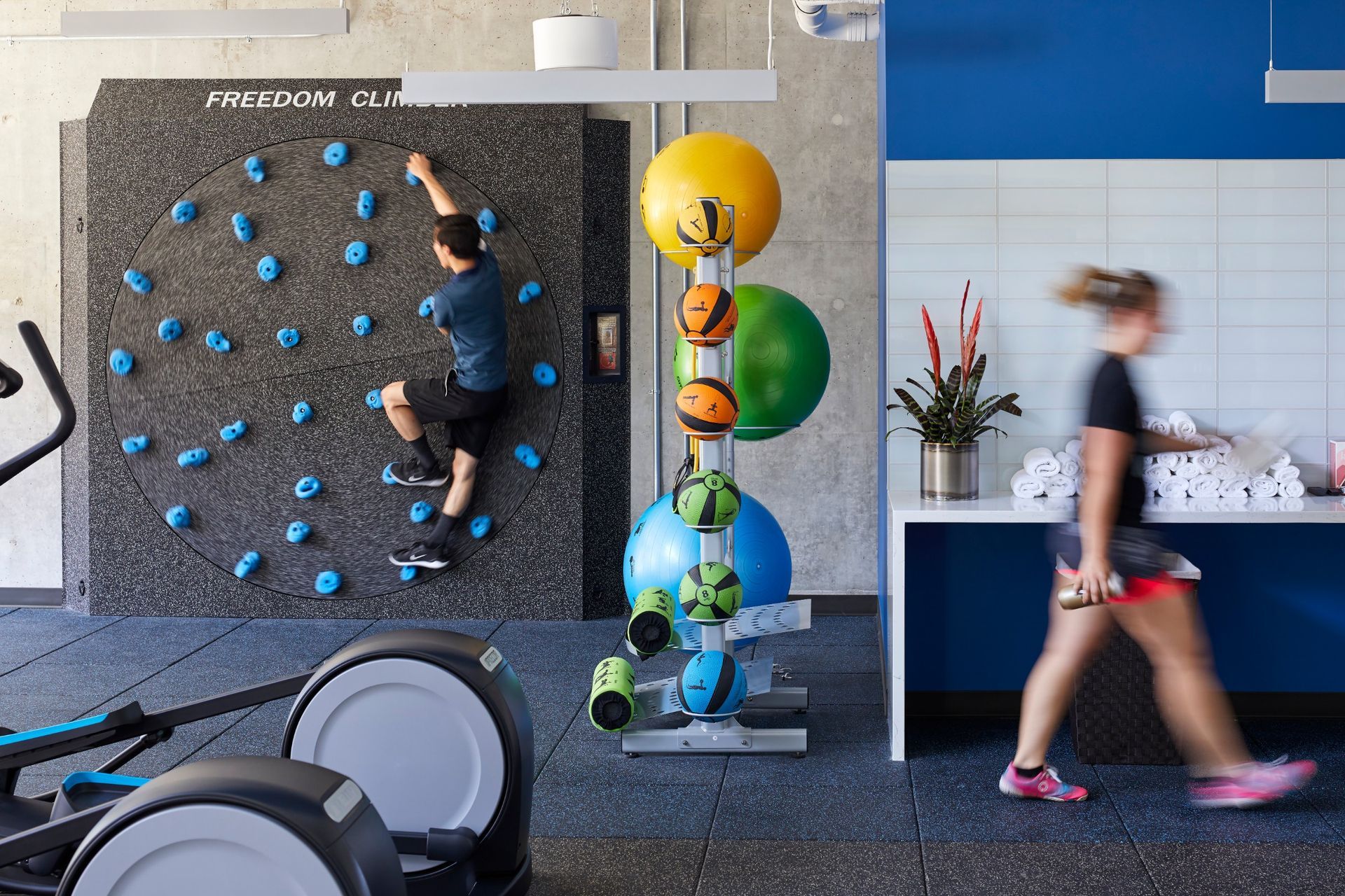 A man is climbing a wall in a gym while a woman walks by.