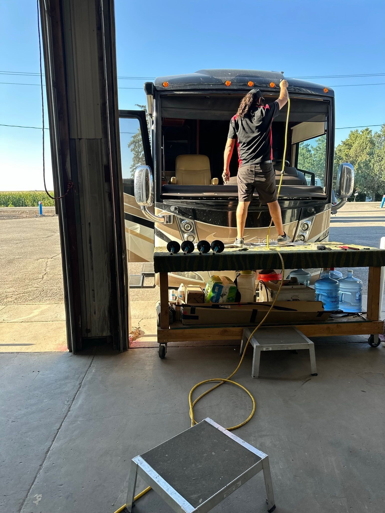A person stands on a workbench cleaning the front windshield of an RV in an open-air garage.