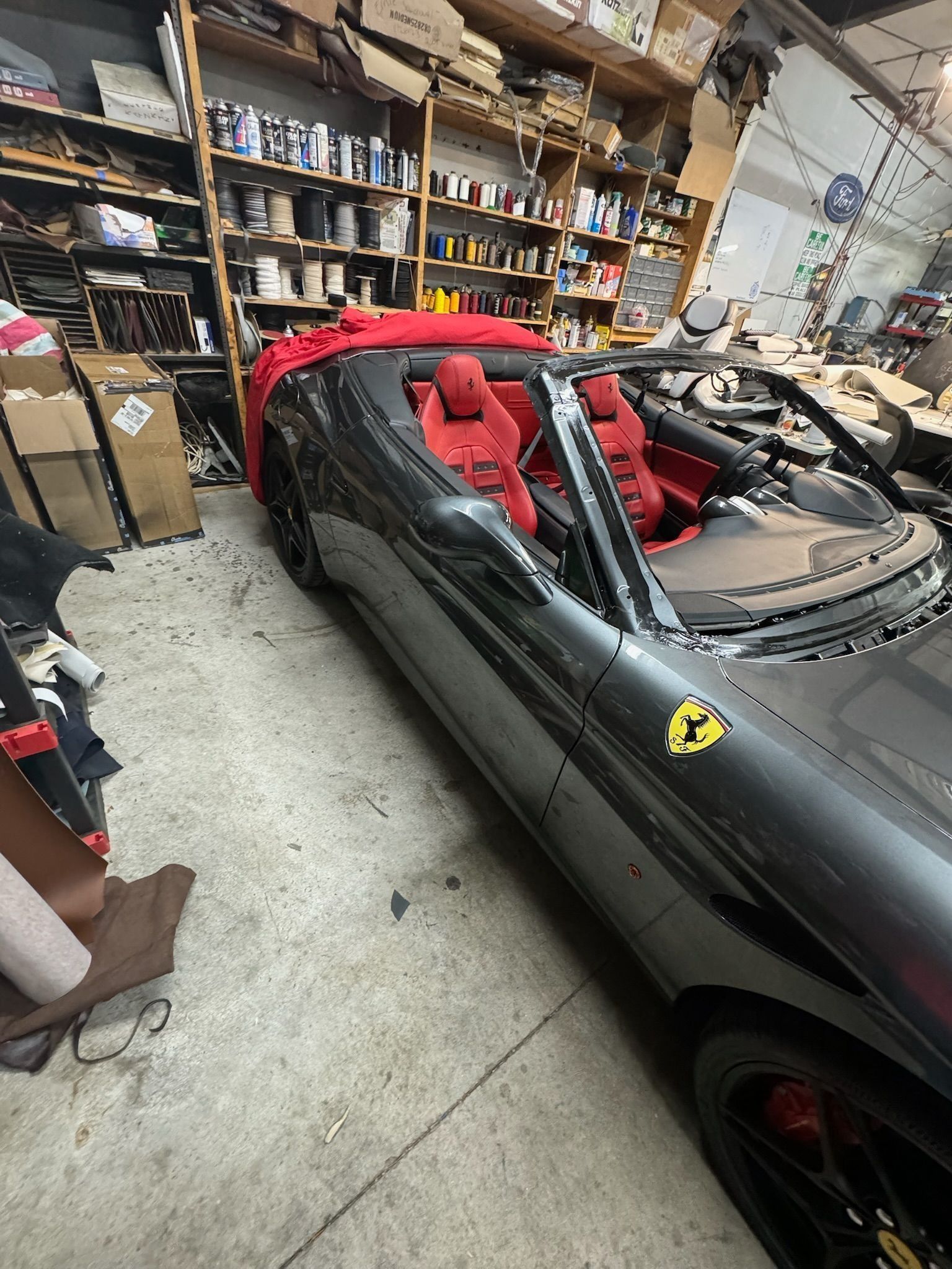 A dark grey Ferrari convertible with red interior parked inside a cluttered workshop with shelves of supplies.