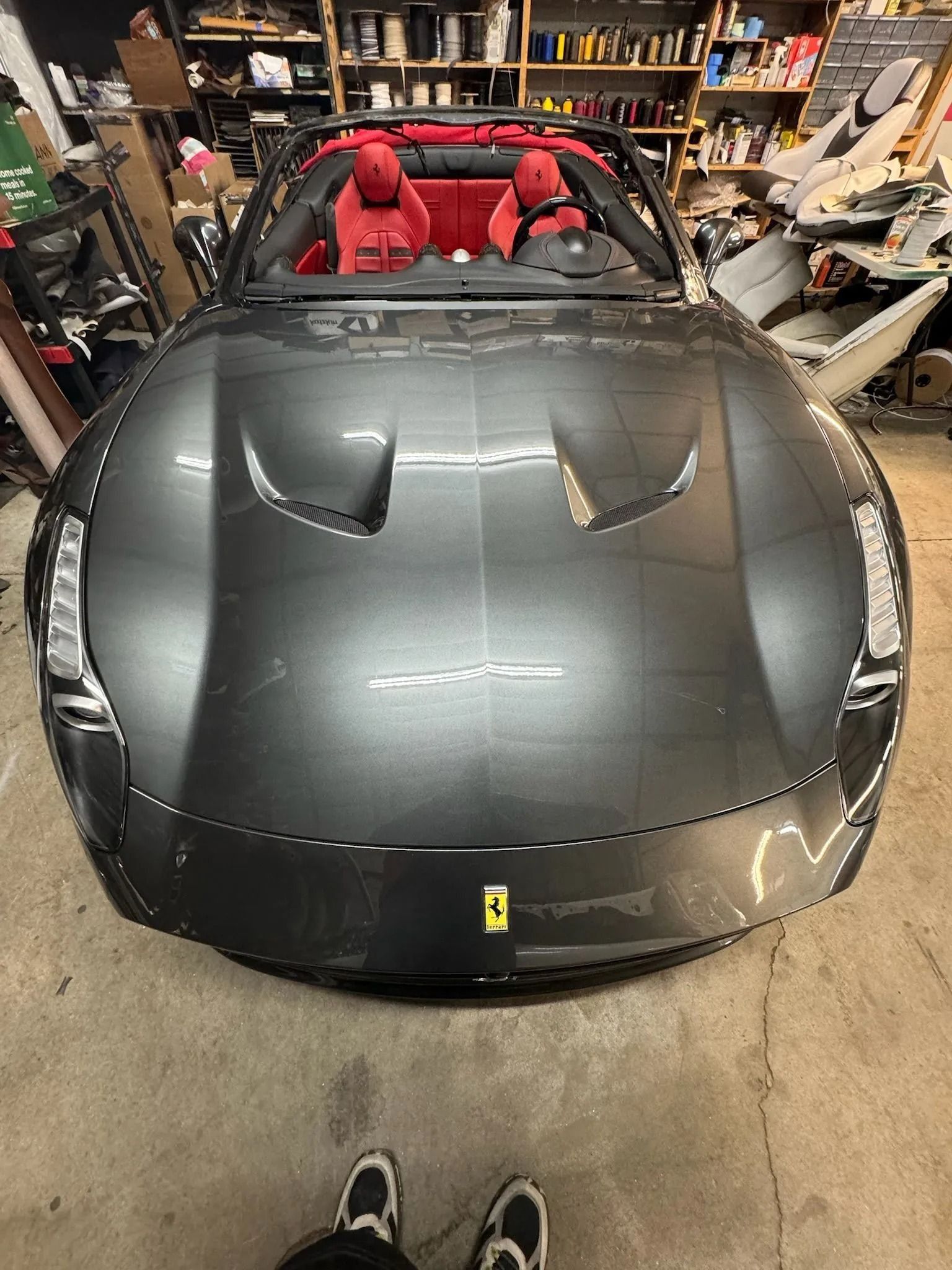 A front-facing, high-angle shot of a metallic grey Ferrari convertible with red interior seats in a garage setting.