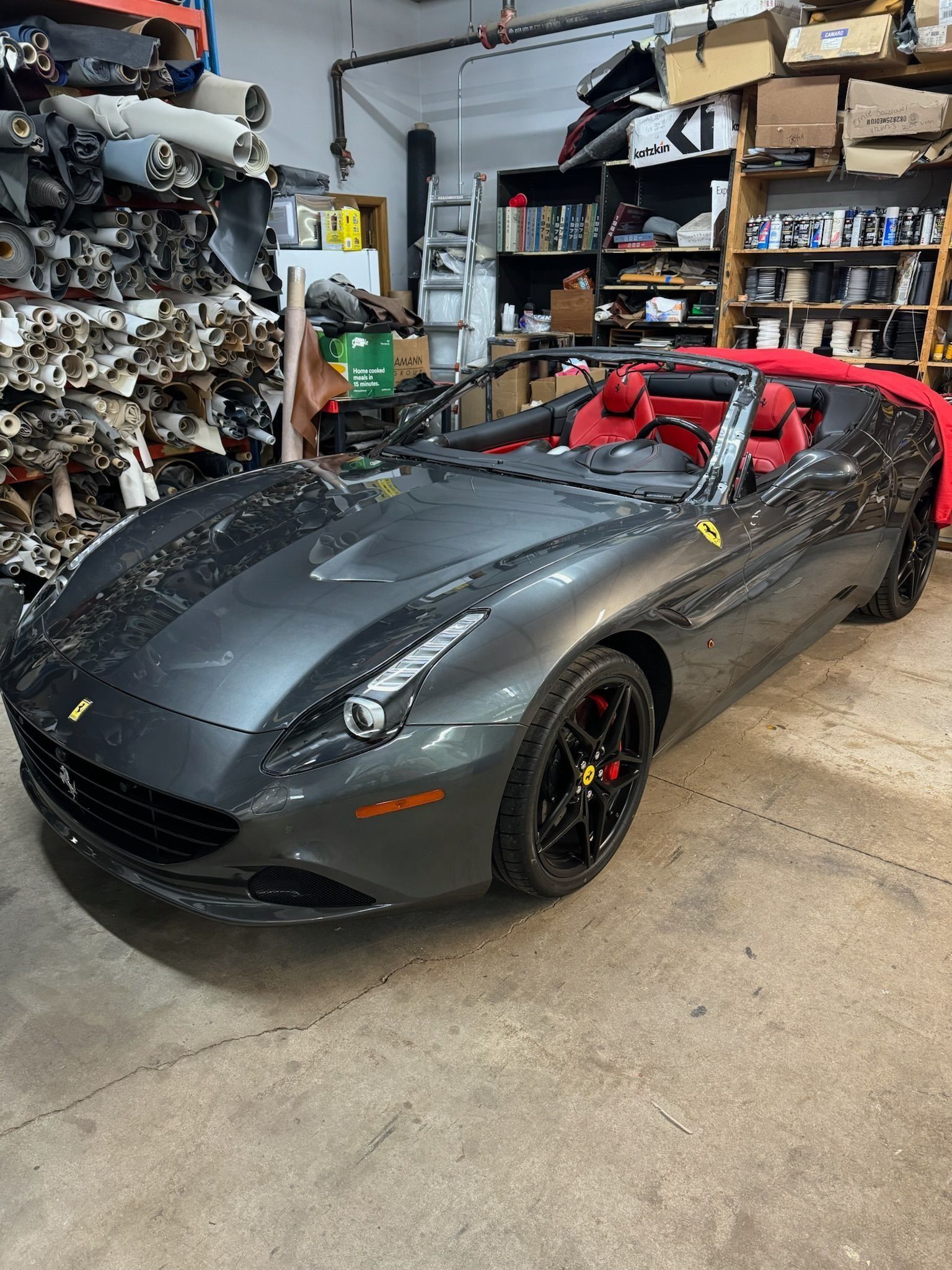 A grey Ferrari convertible with red leather interior parked inside a cluttered industrial garage.