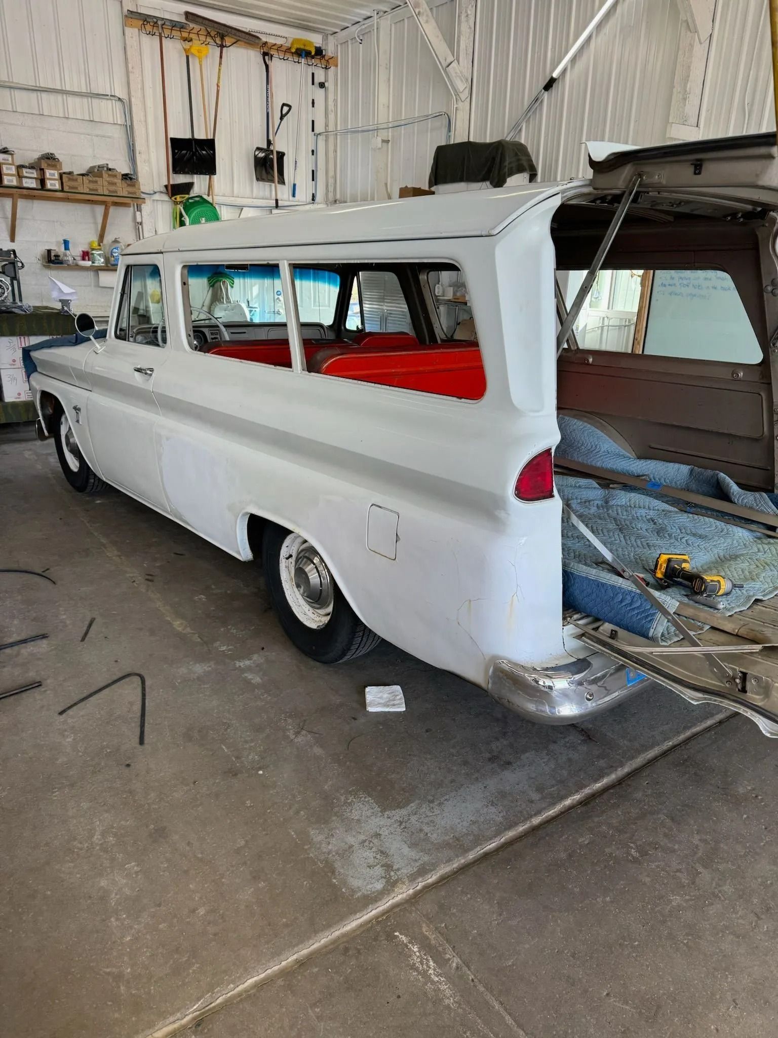 A white vintage Chevrolet Suburban in a garage, undergoing restoration with the rear hatch open and red interior visible.
