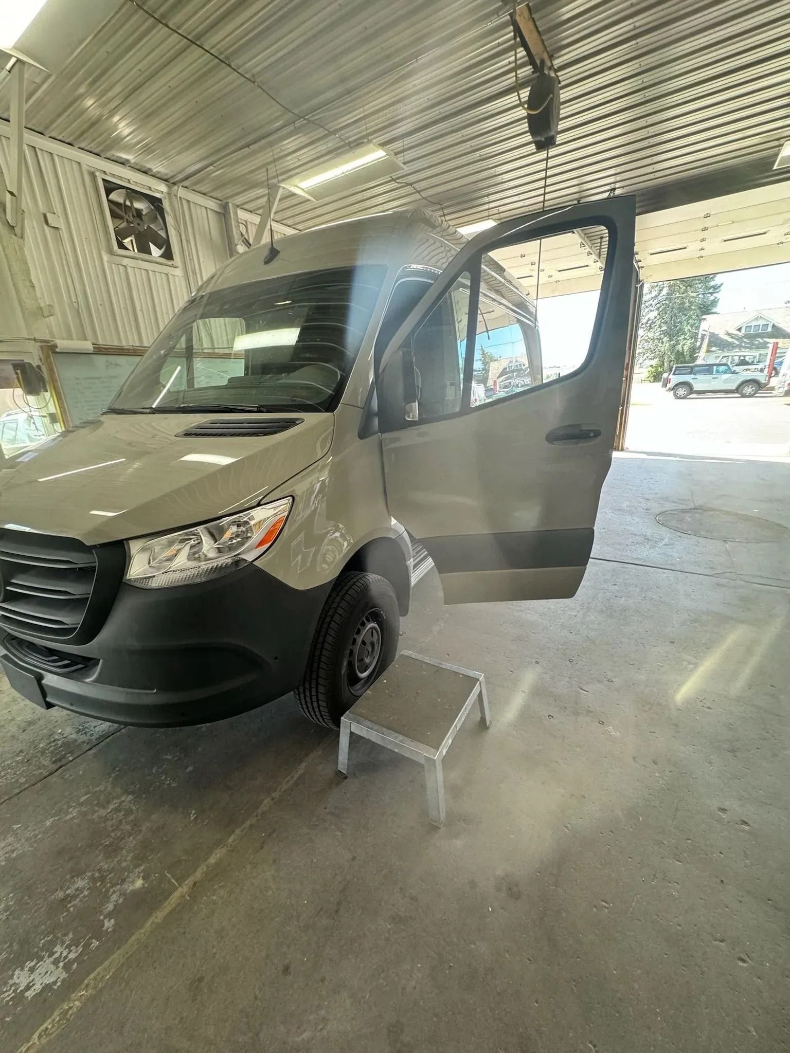 A beige Mercedes-Benz Sprinter van parked inside a workshop with its passenger door open and a metal step stool below.