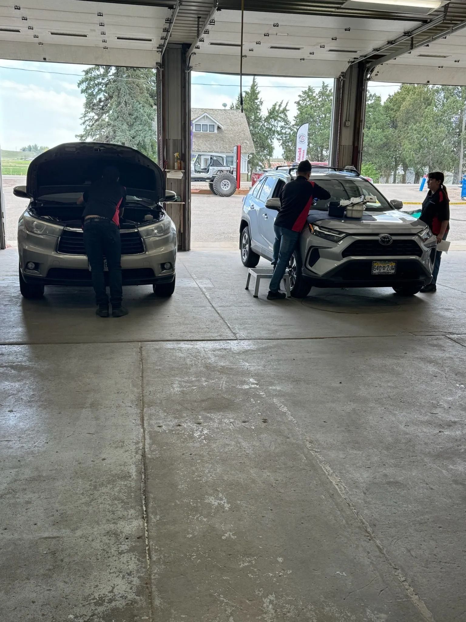 Two mechanics work on two cars in an open-air garage bay; one lifts the hood of a tan car, another checks a white SUV.