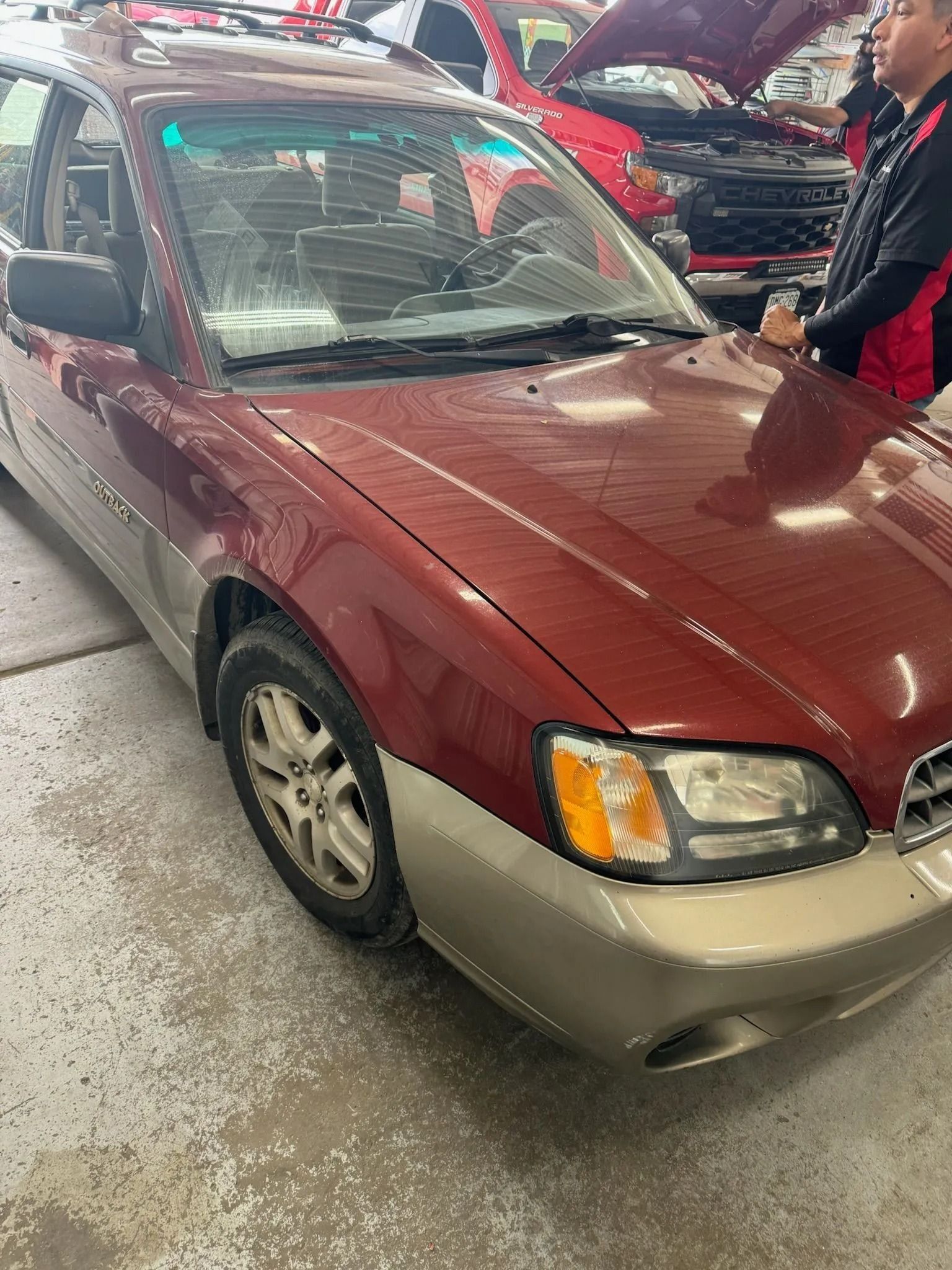 A maroon and tan Subaru station wagon parked in a shop, with a person inspecting a nearby red vehicle in the background.